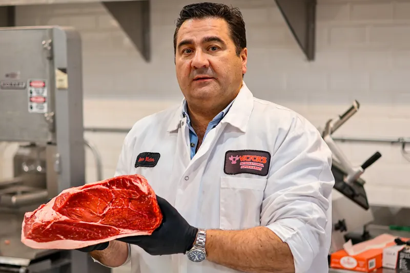 Nuno, wearing a Victor’s International Supermarket coat, holds up a large raw cut of beef in the butcher area