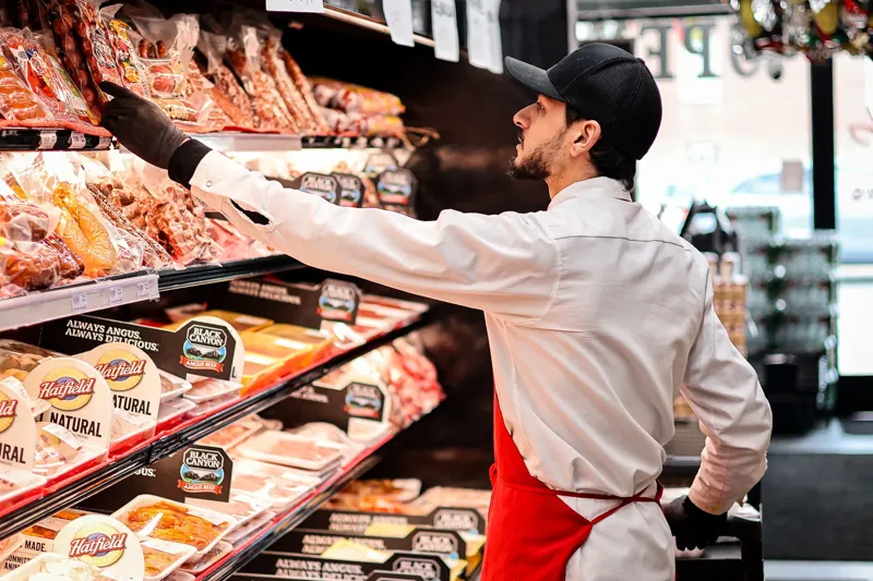 A Victor’s team member in a red apron restocking packaged meats in the refrigerated meat case